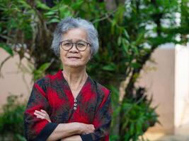 portret van een mooi ouderen Aziatisch vrouw glimlachen en armen gekruiste terwijl staand in een tuin foto