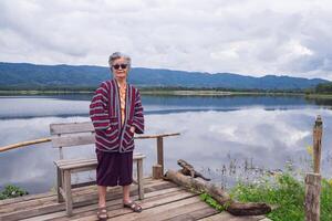 portret van een ouderen Aziatisch vrouw vervelend zonnebril terwijl staand kant de meer. ruimte voor tekst. concept van oud mensen en gezondheidszorg foto