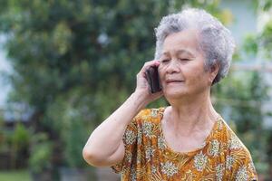 een senior vrouw met kort grijs haar- gebruik makend van een smartphone terwijl staand in een tuin. ruimte voor tekst. concept van oud mensen en communicatie foto