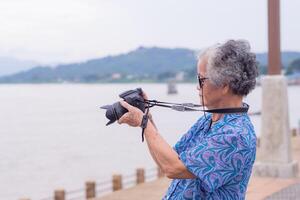 senior vrouw met kort grijs haar- vervelend zonnebril, nemen een foto door een digitaal camera Bij de rivieroever met bergen achtergronden. concept van oud mensen en fotografie