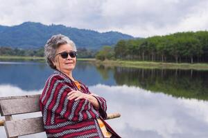 portret van senior Aziatisch vrouw vervelend zonnebril en op zoek weg terwijl zittend Aan een houten stoel kant de vijver. concept van oud mensen en ontspanning foto