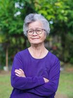 portret van een ouderen vrouw met kort grijs haar, lachend, armen gekruist, en op zoek Bij de camera terwijl staand in een tuin. concept van oud mensen en gezondheidszorg foto