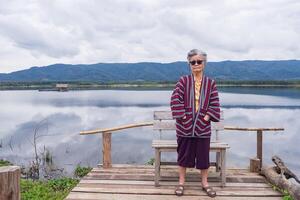 portret van een ouderen Aziatisch vrouw vervelend zonnebril terwijl staand kant de meer. concept van oud mensen en ontspanning foto