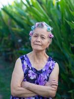 portret van senior vrouw haar- krulspelden, armen gekruiste en op zoek Bij de camera terwijl staand in een tuin. ruimte voor tekst. concepten van oud mensen en gezondheidszorg foto