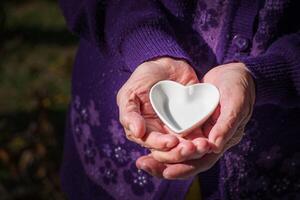 detailopname van een wit keramisch hartvormig Aan palm senior vrouw. Valentijnsdag dag. concept van oud mensen en gezondheidszorg foto