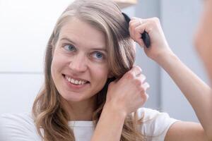 haarverzorging concept. vrouw poetsen haar haar- met kam terwijl staand in de buurt spiegel in badkamer foto