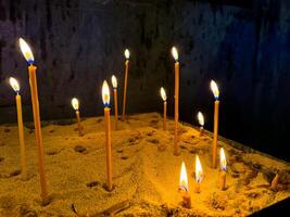 lit kaarsen in zand gevulde kaars houder in kerk tegen donker muur. religieus of geestelijk ceremonie met flikkeren vlammen en warm kaarslicht. brandend kaarsen in kerk. foto