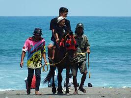 kebumen, centraal Java, Indonesië april 11, 2024 mensen genieten vakantie rijden een paard gedurende helder daglicht Bij ambal strand. zomer familie toerist plek met oceaan Golf achtergrond. foto