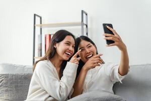 twee Dames zijn zittend Aan een bankstel en nemen een selfie, kijken Bij de slim telefoon, en lachend. foto