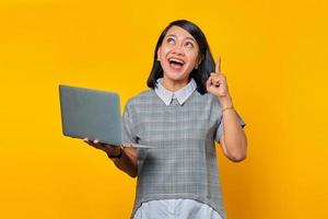 portret van een opgewonden aziatische vrouw die een laptop vasthoudt met een geweldig idee en uitkijkt over een gele achtergrond foto