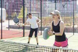 portret van positief jong vrouw en volwassen Mens staand Aan padel tennis rechtbank, Holding racket en bal, glimlachen foto