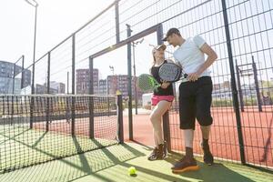 vrolijk atletisch paar lachend gedurende padel tennis bij elkaar passen Aan buitenshuis rechtbank. foto