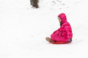 mooi weinig meisje vervelend roze jasje en gebreid hoed wandelen in besneeuwd winter park. kind spelen met ijs het schaatsen bord in Woud. familie vakantie met kind in bergen foto