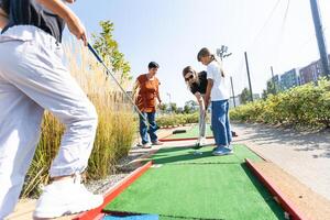 schattig school- meisje spelen mini golf met familie. gelukkig kleuter kind hebben pret met buitenshuis werkzaamheid. zomer sport voor kinderen en volwassenen, buitenshuis. familie vakanties of toevlucht. foto