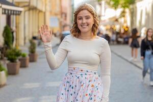 roodharige vrouw glimlachen vriendelijk Bij camera, golvend handen Hallo, hoi, groet of Vaarwel in stad straat foto