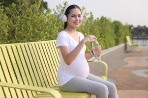 mooi zwanger vrouw drinken fles van zuiver water in de park, gezond en actief zwangerschap levensstijl concept. foto