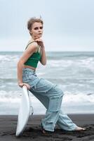 portret van hipster vrouw surfer zittend Aan surfen bord aan het liegen Aan zand, Aan achtergrond van zee golven foto