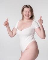 portret van plus grootte vrouw in bodysuit verhogen handen, tonen duimen omhoog, poseren Aan wit achtergrond foto