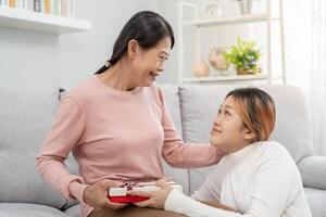 moeder dag, schattig Aziatisch tiener meisje geven geschenk doos naar volwassen midden- leeftijd mamma. liefde, kus, zorg, gelukkig glimlach genieten familie tijd. vieren speciaal gelegenheid, gelukkig verjaardag, gelukkig nieuw jaar, vrolijk Kerstmis foto