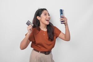 gelukkig geslaagd Aziatisch vrouw vervelend bruin overhemd is Holding haar smartphone en credit kaart over- geïsoleerd wit achtergrond. foto