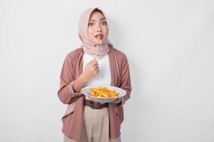 hongerig jong Aziatisch moslim vrouw in hijab Holding vork en bord aan het eten Frans Patat over- geïsoleerd wit achtergrond. foto