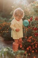 weinig meisje in een geel jurk en rubber laarzen is gieter bloemen in de herfst tuin foto