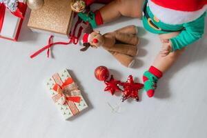 schattig baby in een groen bodysuit en een de kerstman hoed siert de Kerstmis boom met kleurrijk ballonnen foto