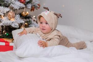 mollig wangedrocht baby in een jumpsuit met hert hoorns leugens Aan een groot wit hoofdkussen onder Kerstmis boom foto