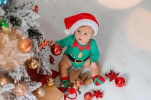 schattig baby in een groen bodysuit en een de kerstman hoed siert de Kerstmis boom met kleurrijk ballonnen foto