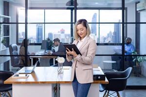 portret van Aziatisch bedrijf Directeur vrouw is staand in kantoor Bij de tafel met digitaal tablet en tonen statistisch tabel tonen jaar- verslag doen van en wolkenkrabber achtergrond foto