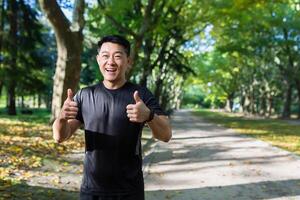 portier van gelukkig geschiktheid trainer in park, Aziatisch Mens glimlachen en op zoek Bij camera, richten vingers omhoog bevestigend, Mens aan het doen sport- in trainingspak Aan zonnig dag in de buurt bomen. foto