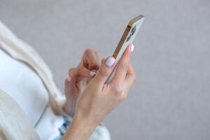 een detailopname foto van een mooi vrouw hand- met een wit manicure is gebruik makend van een goudkleurig telefoon, typen een bericht Aan een smartphone