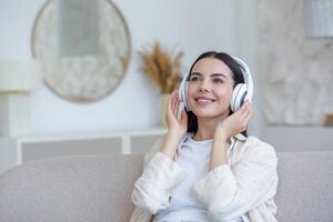 mooi jong vrouw Bij huis ontspannende luisteren naar muziek- van wit hoofdtelefoons zittend Aan de sofa in de dag, het zingen langs en glimlachen blij foto