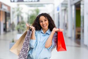 een jong Latijns Amerikaans vrouw toerist kocht souvenirs en cadeaus in een ander stad, land. staand in de boodschappen doen centrum, Holding kleur pakketten, hijs- en tonen hen, glimlacht. foto