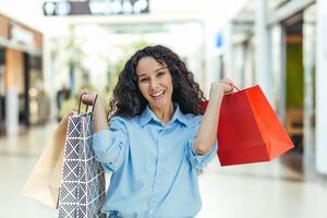 een jong Latijns Amerikaans vrouw toerist kocht souvenirs en cadeaus in een ander stad, land. staand in de boodschappen doen centrum, Holding kleur pakketten, hijs- en tonen hen, glimlacht. foto