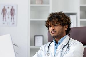 detailopname foto. portret van een jong Indisch mannetje dokter zittend in een kantoor in een wit jas en werken afgeleid Bij een laptop. foto