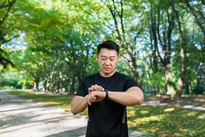 actief levensstijl, modern technologieën. jong sportief Aziatisch Mens toepassingen een geschiktheid armband, looks Bij een slim kijk maar terwijl jogging buiten in de park. maatregelen hart tarief, aantal van stappen, afstand. foto