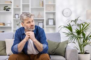 volwassen volwassen Mens nadenkend zittend alleen Bij huis Aan sofa dichtbij omhoog, grijs haren persoon denkt looks naar venster, depressief verdrietig. foto