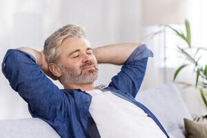 detailopname foto. senior grijs haar Mens zit Aan de sofa Bij huis en rust, duurt een dutje. hij zetten zijn handen achter zijn hoofd, Gesloten zijn ogen, ontspannen na een moeilijk dag werk. foto