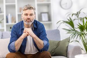 portret van een eng knap grijs haar Mens in een blauw overhemd zittend Aan de sofa Bij huis en ernstig op zoek Bij de camera, kruispunt zijn armen in voorkant van hem. foto