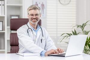 portret van een volwassen volwassen grijs haar dokter binnen een medisch kantoor, de dokter is glimlachen en op zoek Bij de camera, de Mens is zittend Bij een tafel met een laptop en in een wit medisch jas. foto