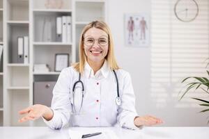 portret van een vrouw dokter zittend in de kantoor Bij de tafel in een wit jas en met een stethoscoop, pratend en overleg plegen online Aan camera. hij glimlacht, voert een medisch webinar, opleiding. foto