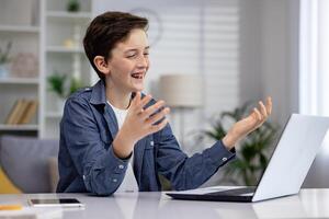 kind jongen, tiener zit Bij huis Bij de tafel in voorkant van de laptop. hij studie, praat Aan een telefoontje met vrienden, glimlacht, gebaren met zijn handen. foto