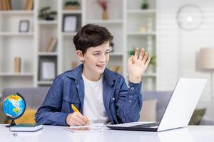 een schattig leerling jongen zit in voorkant van een laptop Bij een tafel in een helder kamer Bij huis, studies van een afstand, looks Bij de computer scherm, golven in groet, schrijft in een notebook. foto