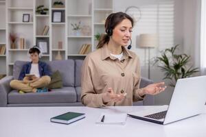 zakenvrouw in koptelefoon zittend Bij tafel in leven kamer in voorkant van laptop, hebben telefoongesprek, aantrekkelijk vrouw werken in telefoontje centrum, werken van huis, moeder werken met kind in achtergrond. foto