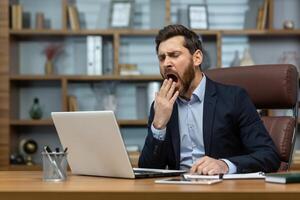 volwassen Mens geeuwen binnen klassiek kantoor, senior zakenman in werkplaats slapeloos en overwerkt, baas in bedrijf pak zittend Bij bureau, gebruik makend van laptop Bij werk. foto