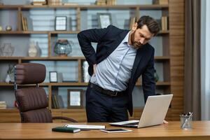 volwassen Mens werken in modern kantoor met laptop, senior baas hebben erge, ernstige terug pijn, zakenman Bij werkplaats overwerkt werken laat masseren zijn terug met hand, erge, ernstige pijn van zitten. foto