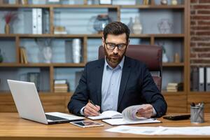 echt en gefocust financier accountant Aan papier werk binnen kantoor, volwassen Mens gebruik makend van rekenmachine en laptop voor berekenen rapporten en samenvatten rekeningen, zakenman Bij werk. foto