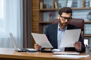 echt en gefocust financier accountant Aan papier werk binnen kantoor, volwassen Mens gebruik makend van rekenmachine en laptop voor berekenen rapporten en samenvatten rekeningen, zakenman Bij werk . foto