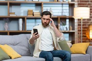 verdrietig en depressief Mens alleen Bij huis zittend Aan bank, freelancer in gewoontjes kleren Holding smartphone, ontevreden lezing slecht nieuws online van telefoon. foto